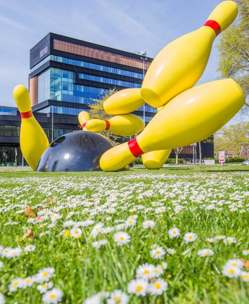The Flying Pins Monument sculpture in Eindhoven — giant yellow bowling pins toppling over a black ball, with daisy flowers in the foreground and a modern building behind